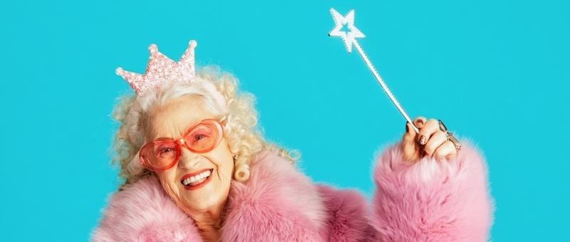 Beautiful smiling senior old woman with white curly hair in fairy fancy dress wearing pink fur coat, star-shaped glasses, and holding a star-shaped wand against pink background.